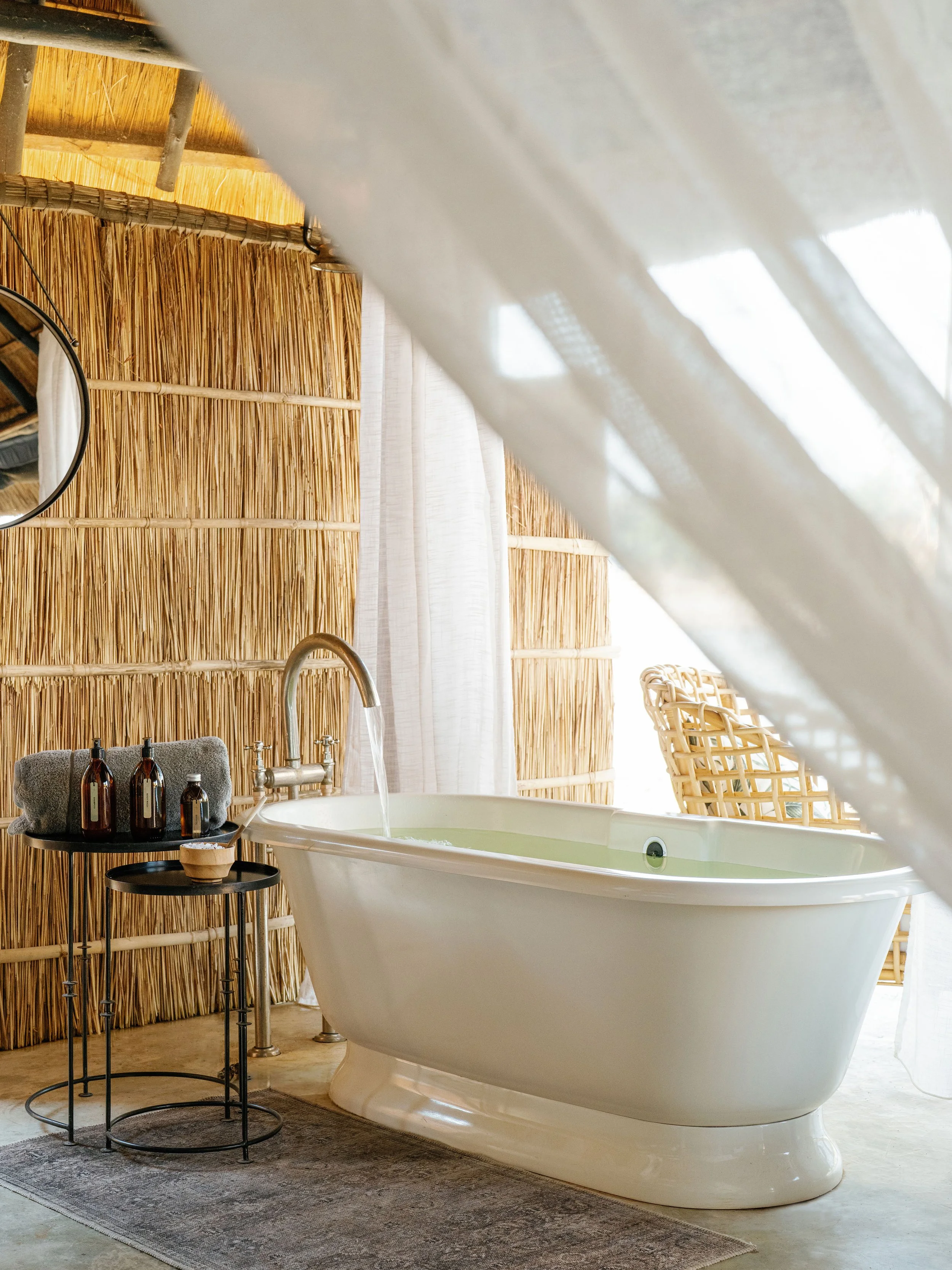 A white freestanding bathtub filled with water in a bathroom with natural light, a bamboo wall, and wicker chairs in the background.