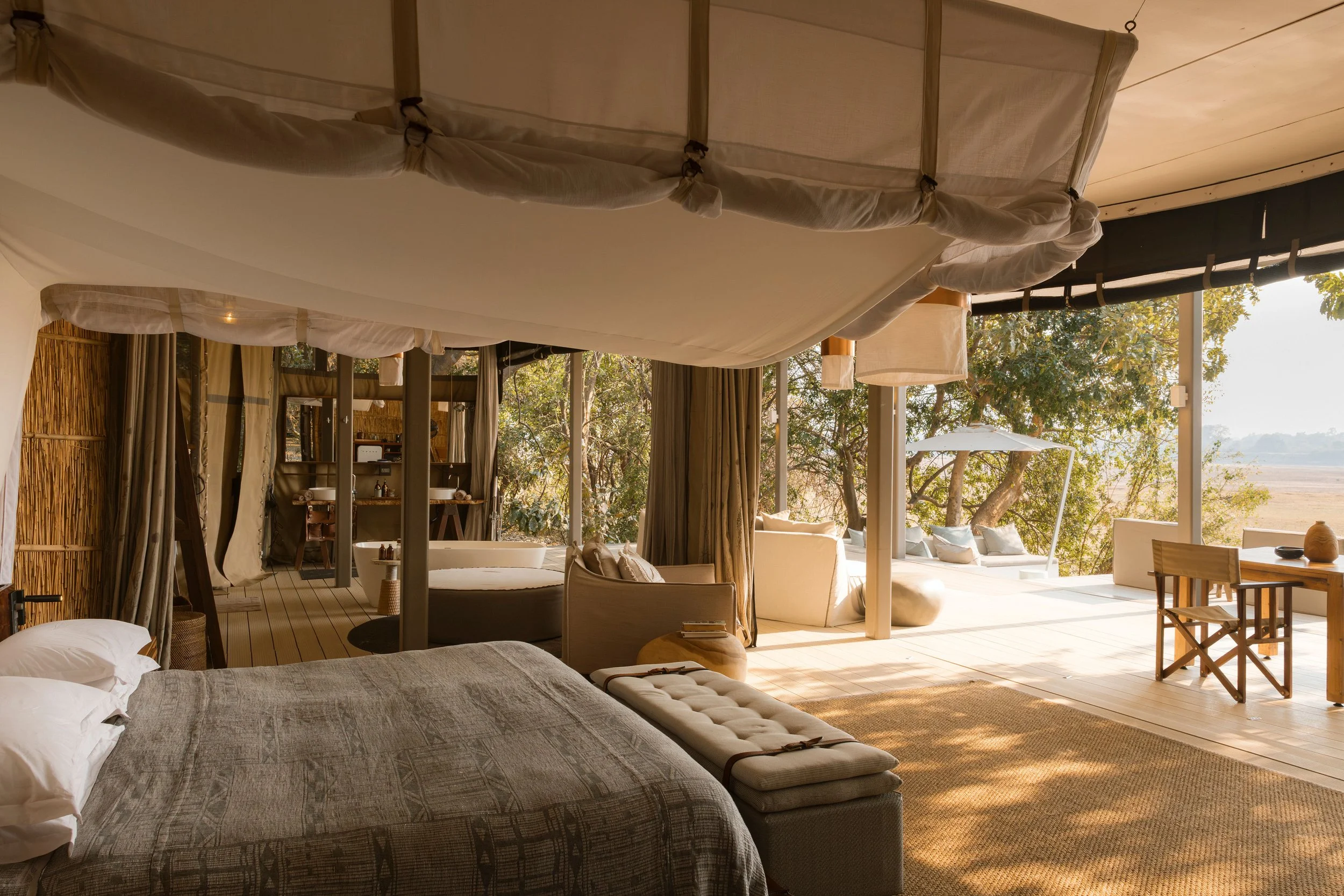 A spacious bedroom with a large bed, beige bedding, and pillows in the foreground. The room features an open design with large sliding glass doors that lead to an outdoor area with trees and a scenic landscape. There are outdoor seating arrangements,
