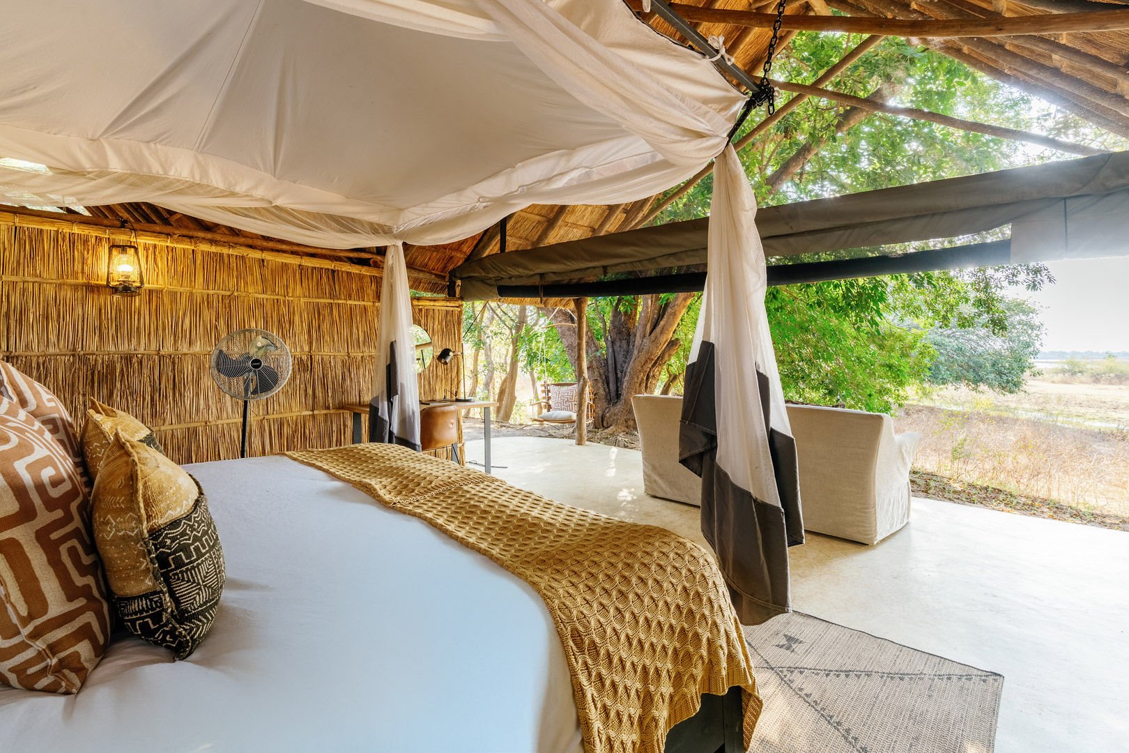 A cozy outdoor bedroom with a canopy-style roof, a large bed with decorative pillows and a yellow blanket, and a fan. There is a desk with a chair and a reading lamp, and outdoor seating with views of trees and open land.