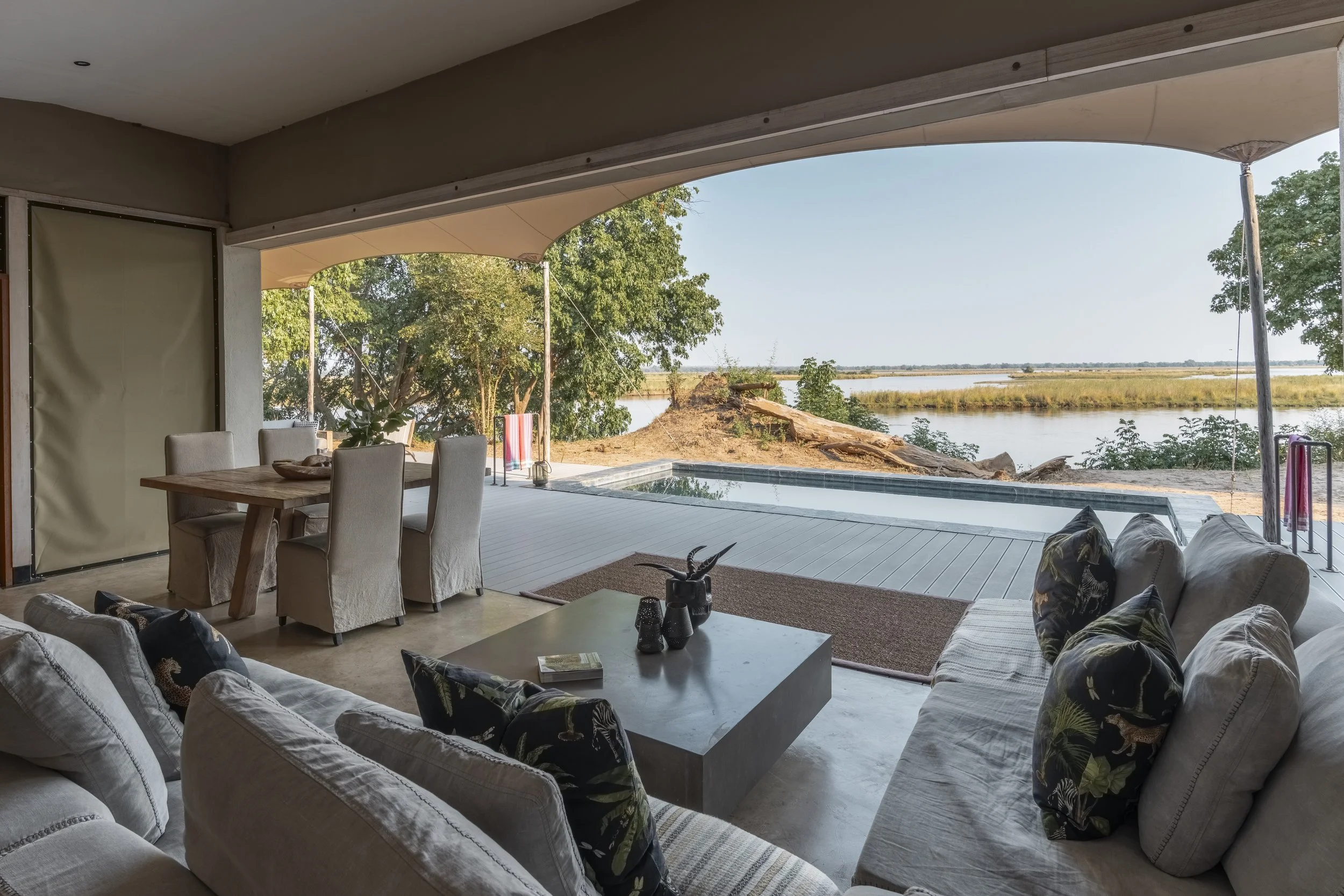 Living room with beige sofas, a coffee table with decorative vases, and a view of a river and trees outside through an open wall with a patio, under a curved canopy.