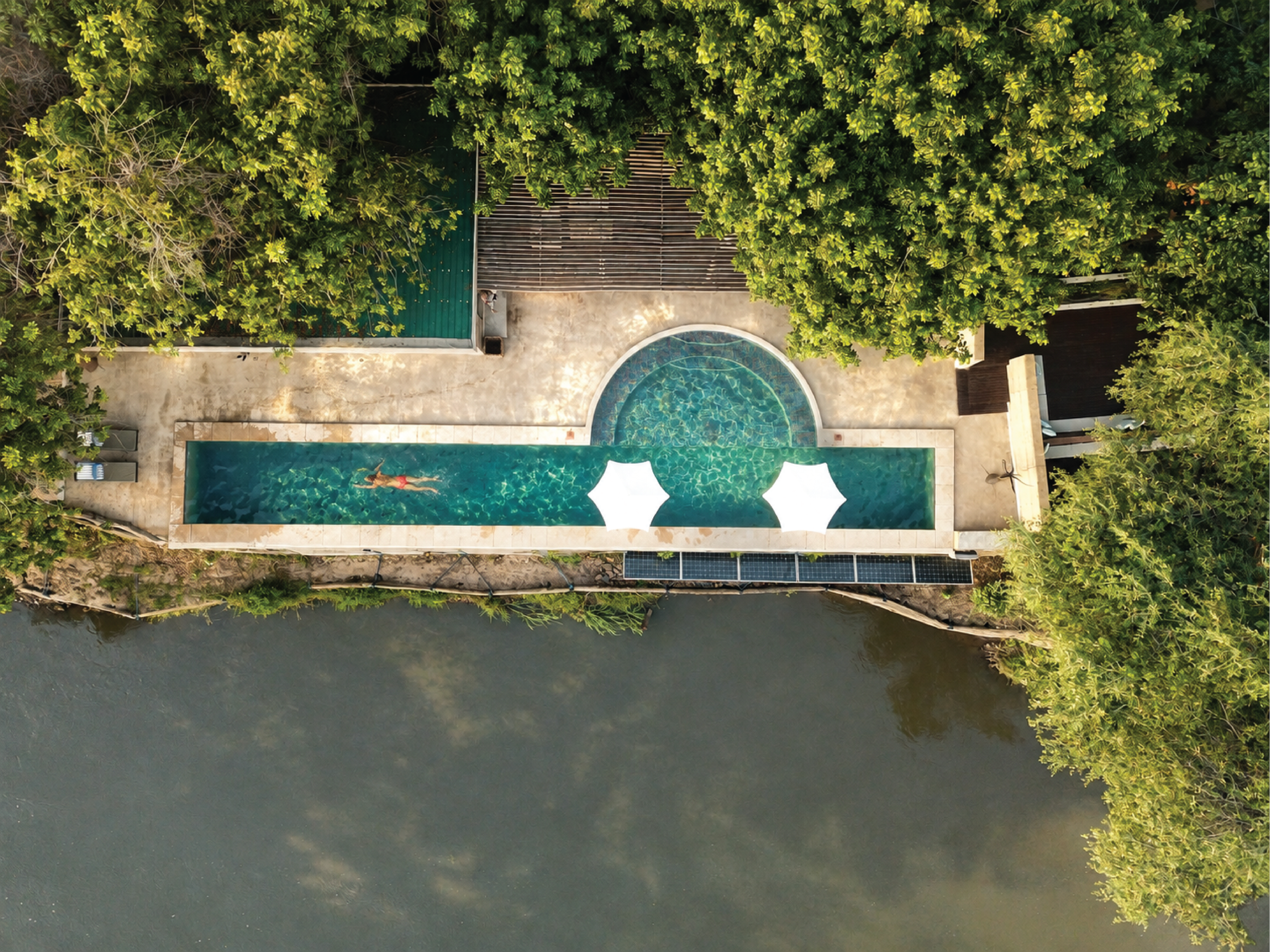 Aerial view of a swimming pool with a person swimming, surrounded by many green trees and a river running alongside.