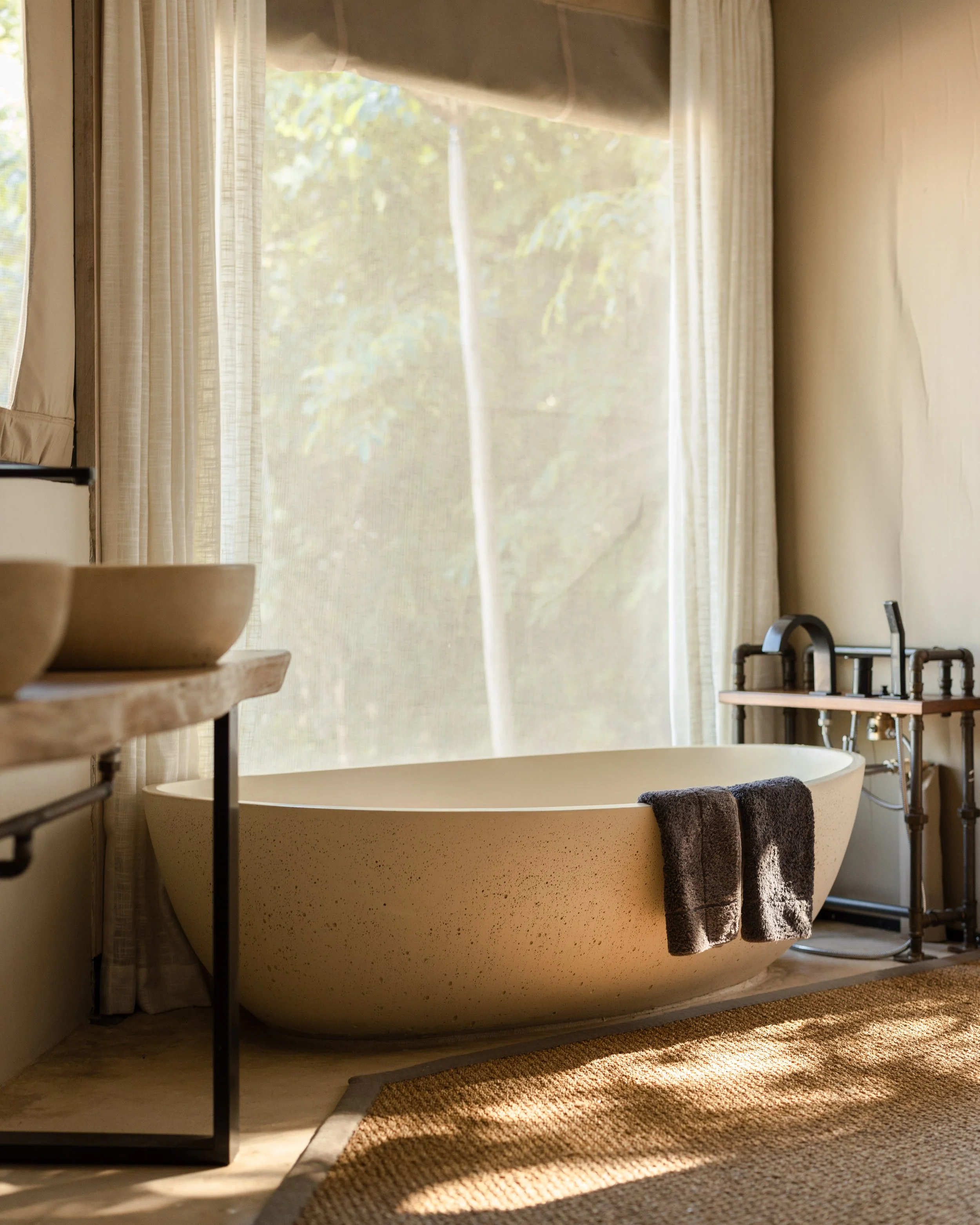 A bathroom with a large freestanding bathtub, a brown towel hanging over the edge, and a window with sheer curtains letting in natural light. There is a rug on the floor and some pipes with a faucet on the right side.
