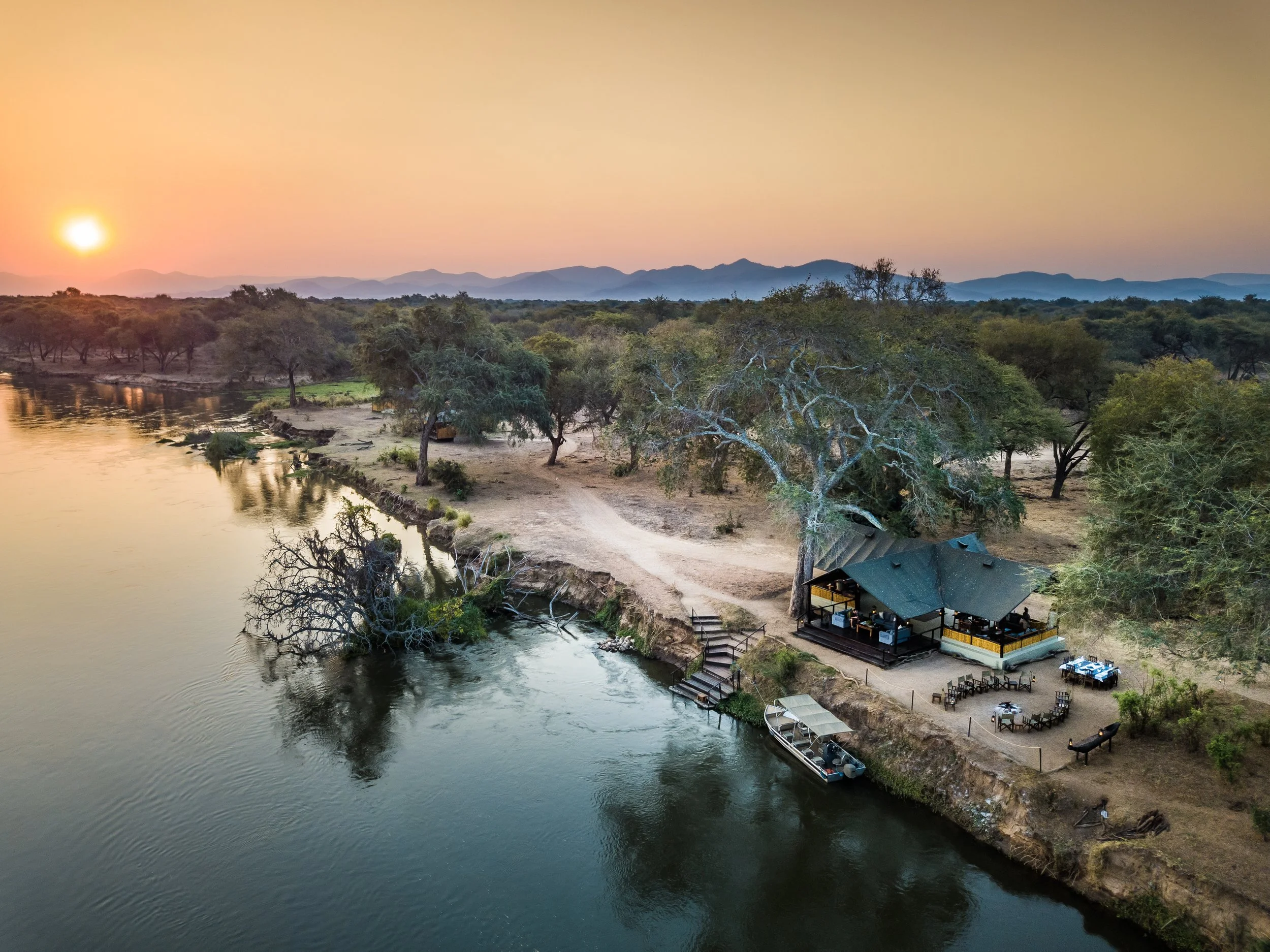 A river flowing past a sandy beach with trees, and a building with outdoor seating on the shore at sunset, with mountains in the background.