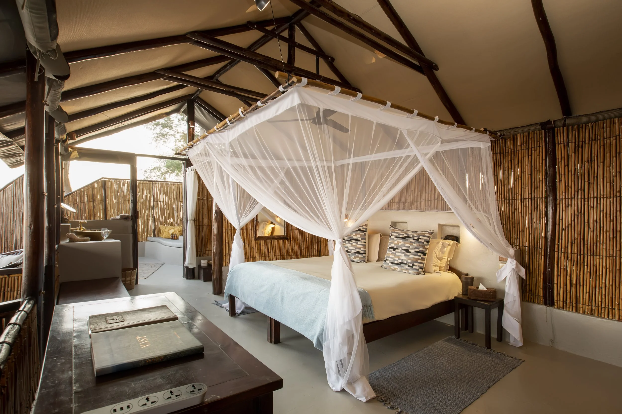A bedroom with a four-poster bed draped with white mosquito netting, wooden furniture, and bamboo wall accents, illuminated by soft natural light.
