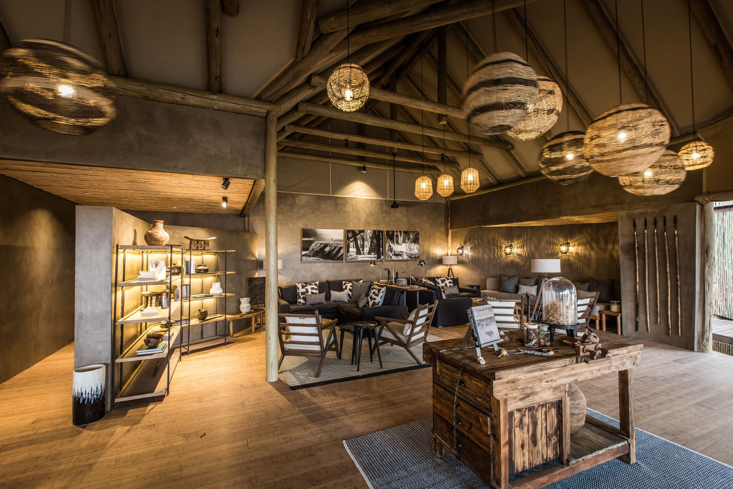 Interior of a rustic lounge with wooden flooring, exposed wooden beams on the ceiling, and modern lighting fixtures made of woven material. The room has a seating area with sofas, chairs, and artwork on the walls, creating a cozy atmosphere.
