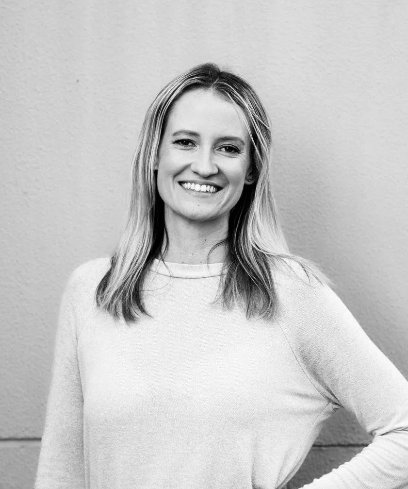 Black and white portrait of a smiling woman in a light-colored sweater standing against a plain wall.