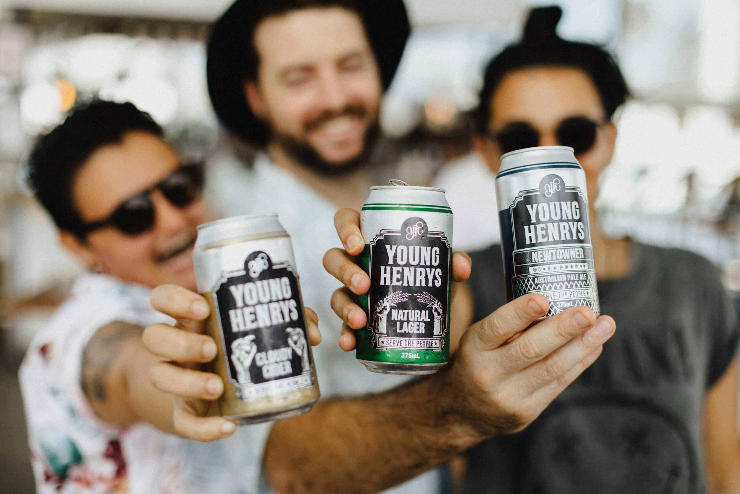 Three friends holding up Young Henrys beer cans towards the camera, smiling and celebrating. A candid moment of brand love and authentic community connection.