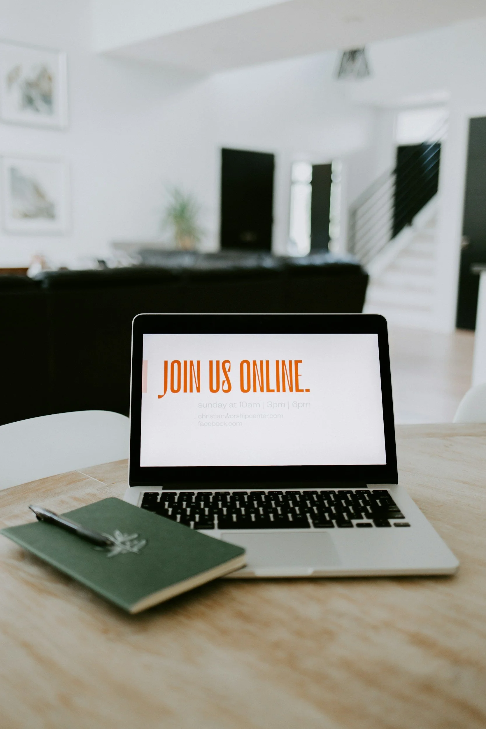 A laptop on a wooden table displaying a message to join an online event, with a notebook and pen next to it in a bright, modern home interior.