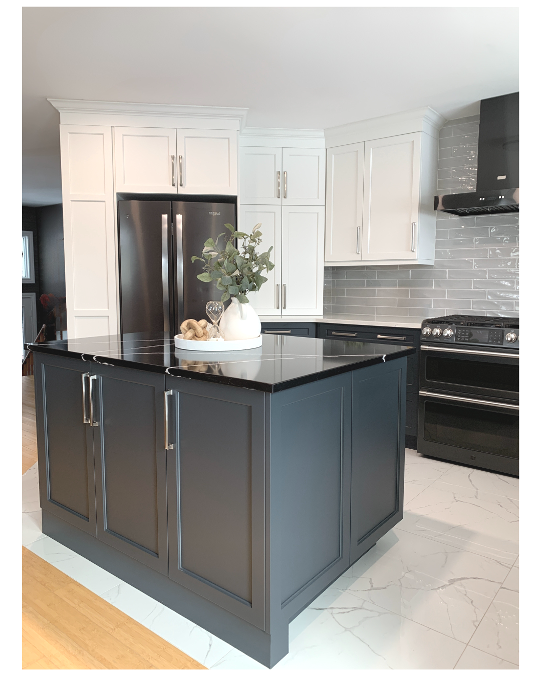 Modern kitchen with white upper cabinets, dark lower cabinets, black countertops, stainless steel appliances, light gray tiled backsplash, and a black island with decorative items on top.