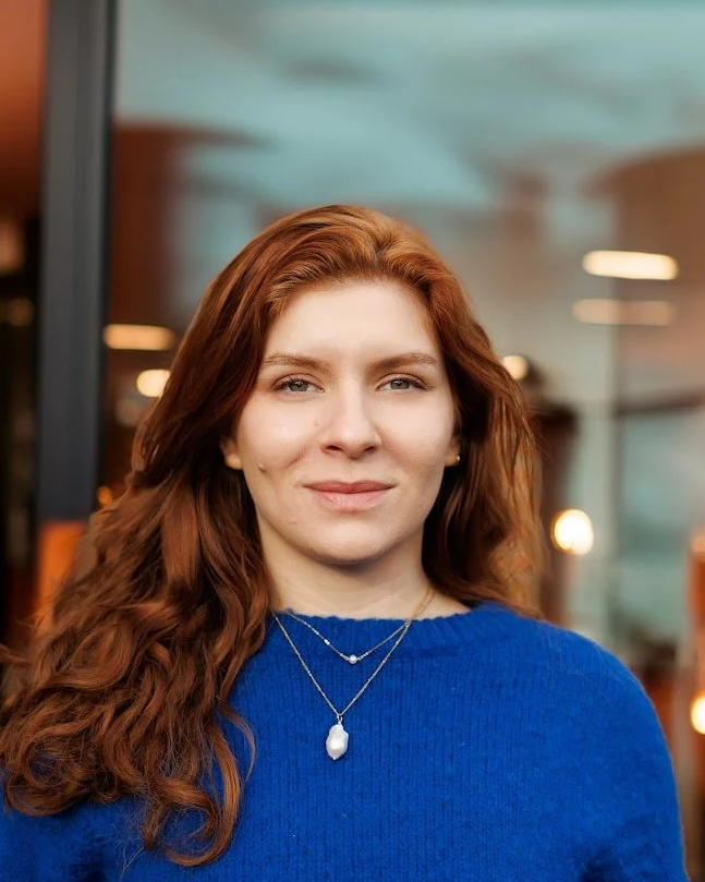 A young woman with long red hair, wearing a royal blue sweater and layered necklaces, standing indoors with blurred lights in the background.