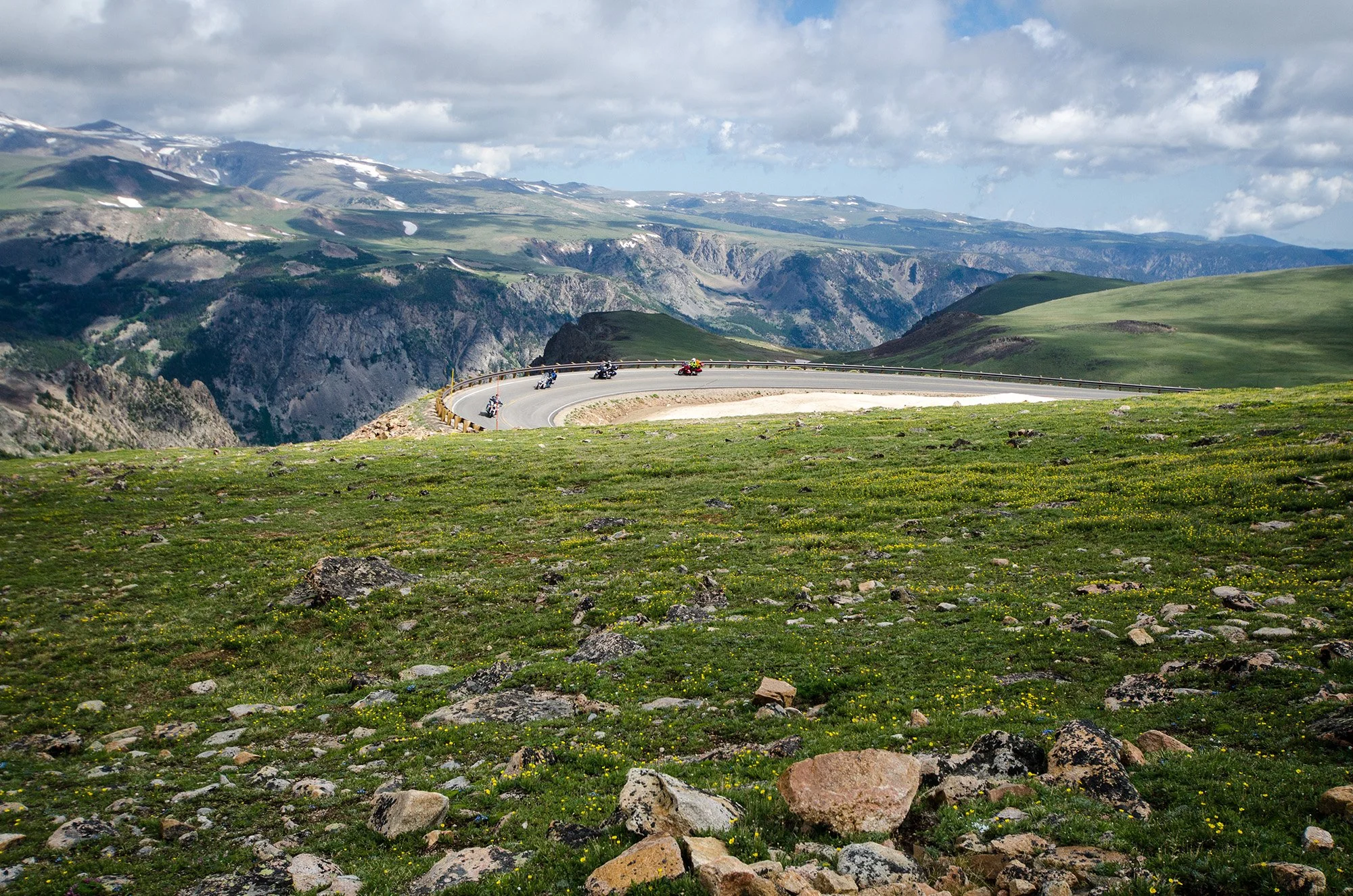 Motorcycles riding on a winding mountain road with green grassy hills, rocky terrain, and snow-capped mountains in the distance under cloudy sky.
