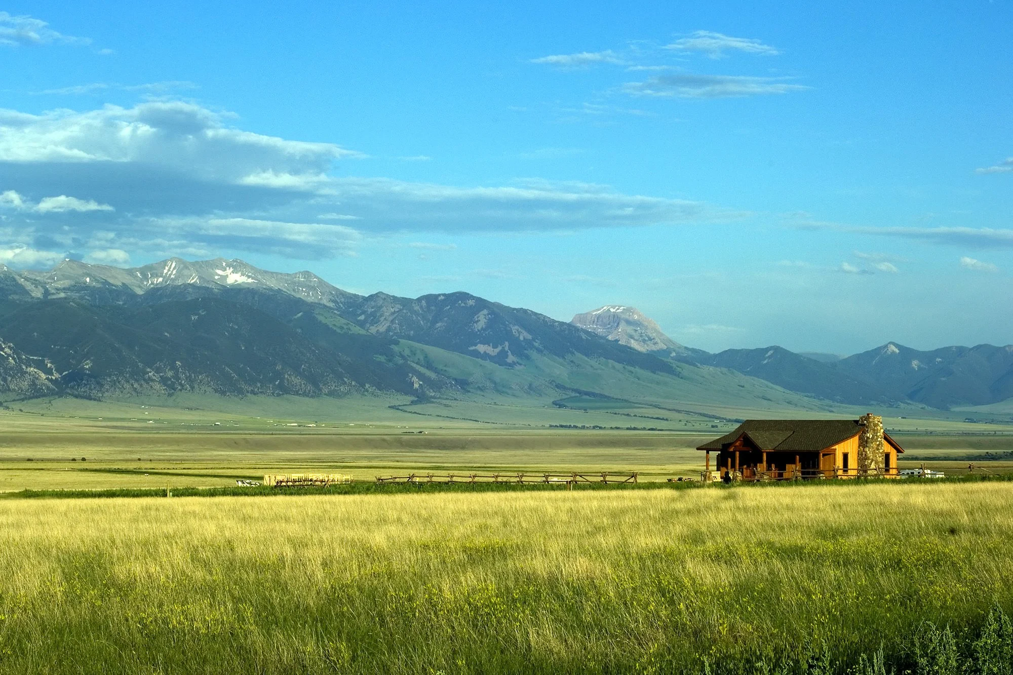 A scenic landscape featuring green fields in the foreground, a small wooden house with a chimney on the right, and a mountain range with snow patches in the background under a partly cloudy blue sky.