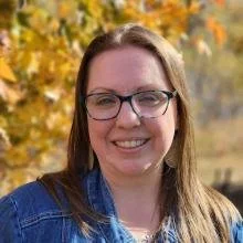 A woman with glasses smiling outdoors during fall, with colorful leaves in the background.