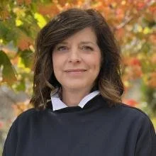 A woman with shoulder-length dark hair wearing a navy blue top standing outdoors with fall foliage in the background.