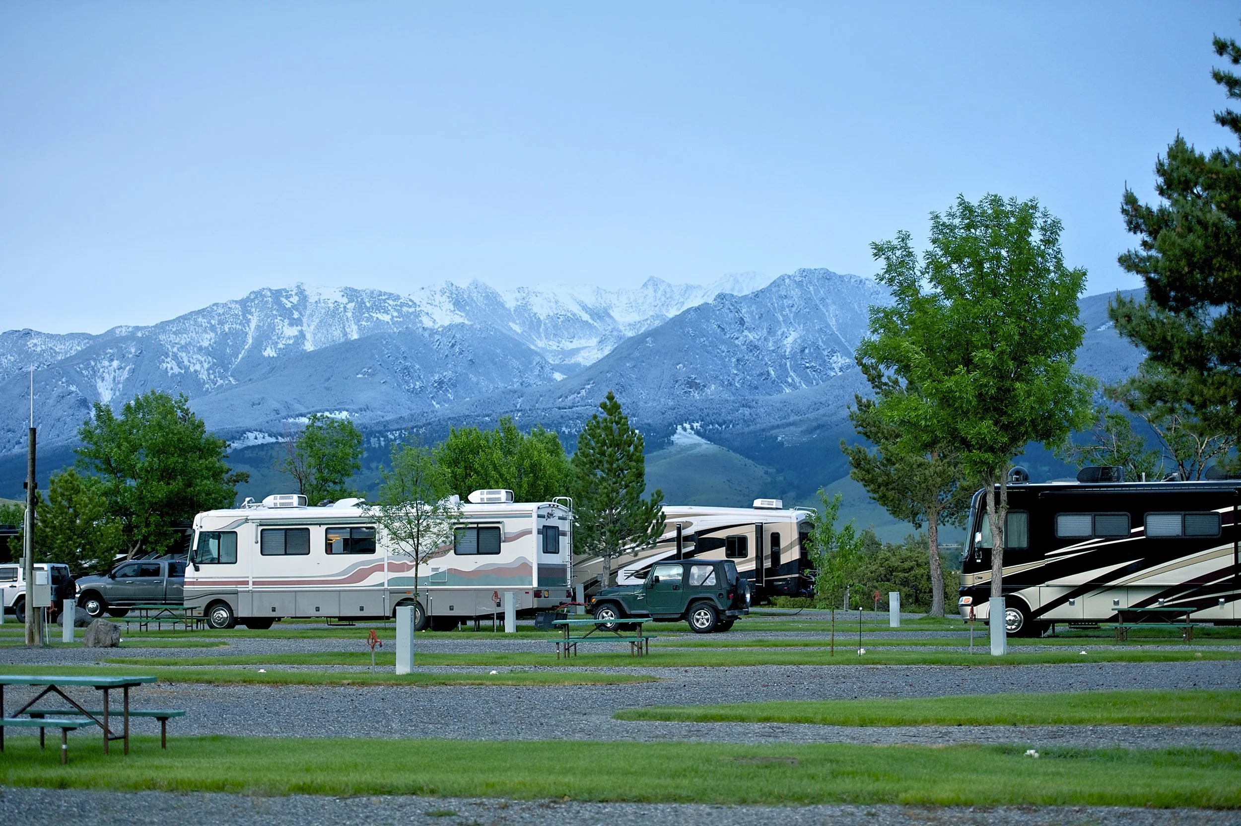 A scenic view of a mountain range with snow-capped peaks in the background and green trees in the foreground. There are RVs and vehicles parked in a campground setup below.