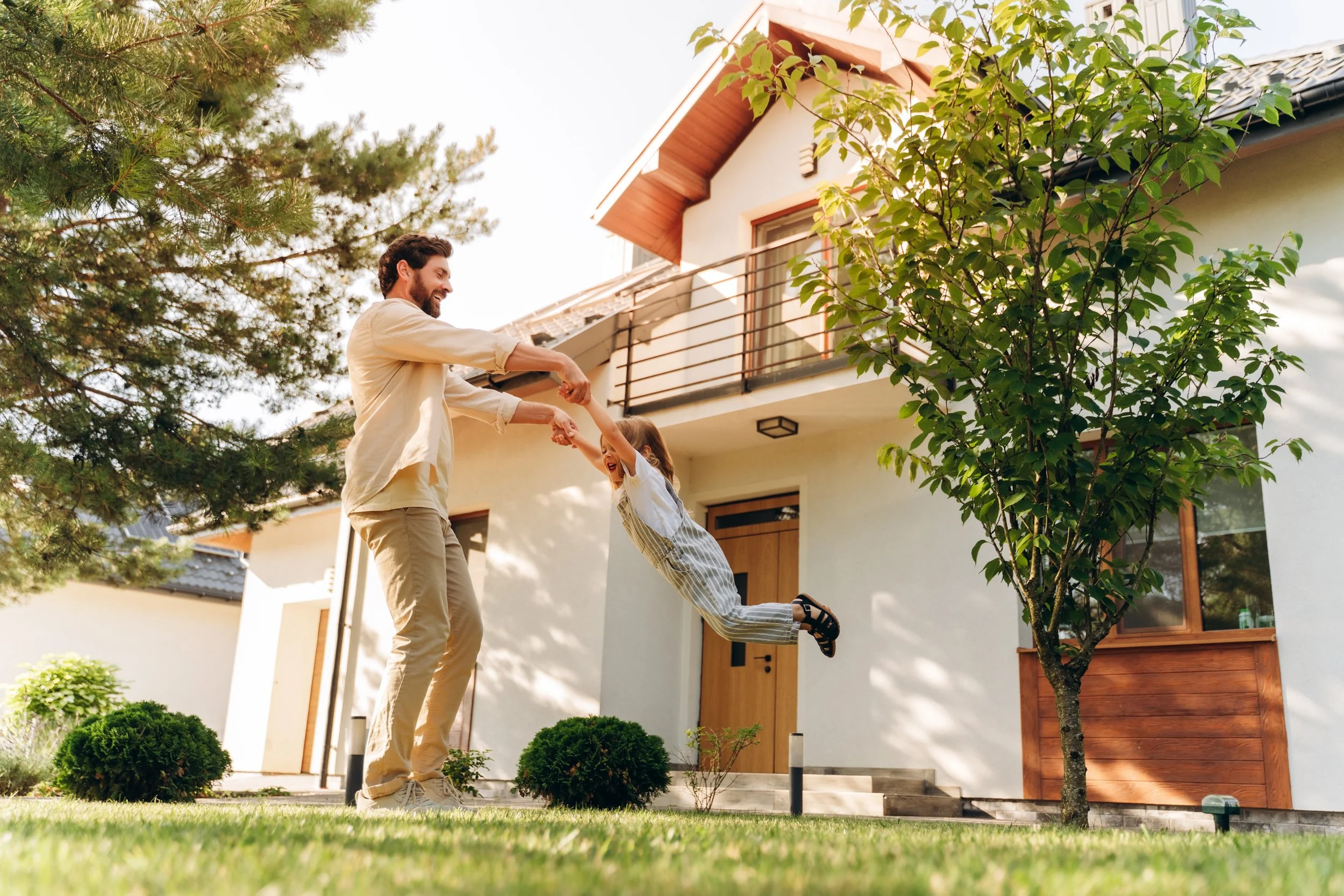 A man and a girl are playing outside in front of a modern house with a balcony and a wooden door. The girl is flying through the air while holding the man's hands. There are trees and bushes around them, and the sky is clear.