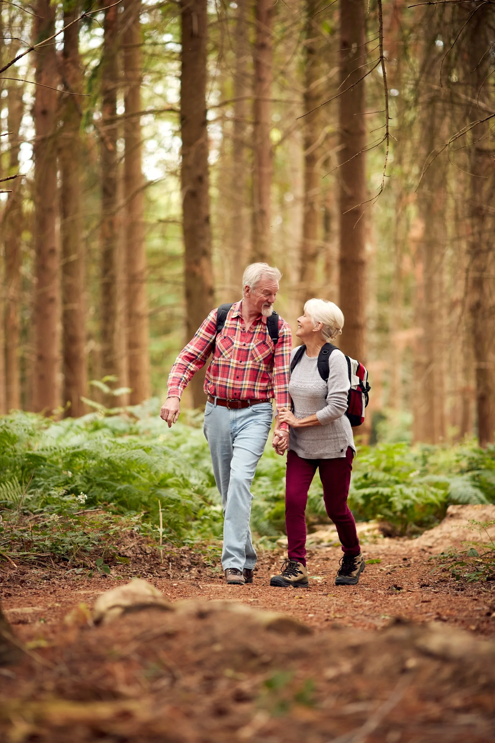 An elderly couple hiking together in a forest, holding hands and smiling at each other, surrounded by tall trees and green foliage.