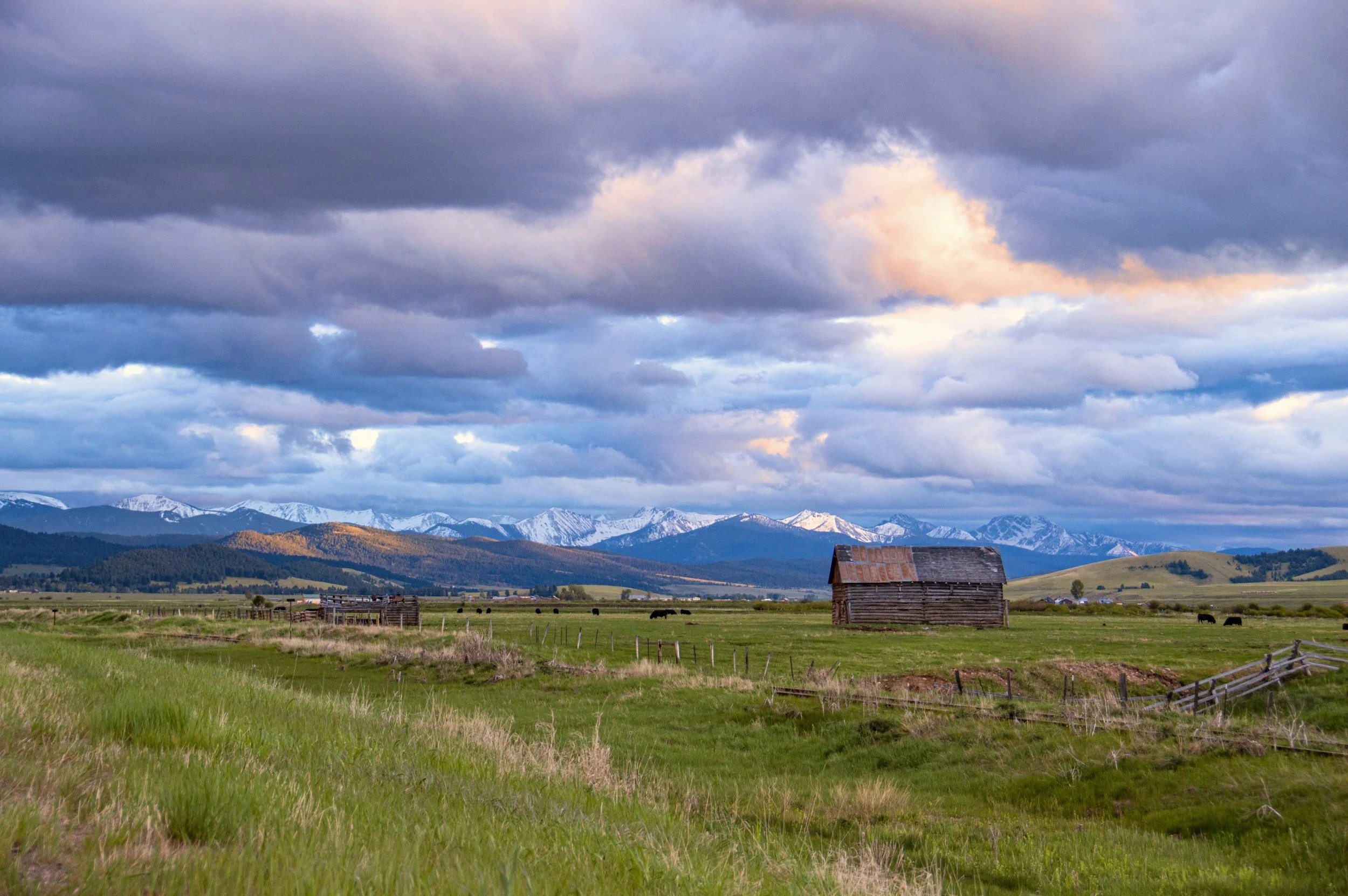 A rural landscape with green fields and a rustic wooden barn, snow-capped mountains in the background, and a cloudy sky with hints of pink and purple at sunset.