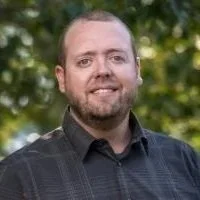 A man with short hair and a beard wearing a dark collared shirt, standing outdoors with trees in the background.