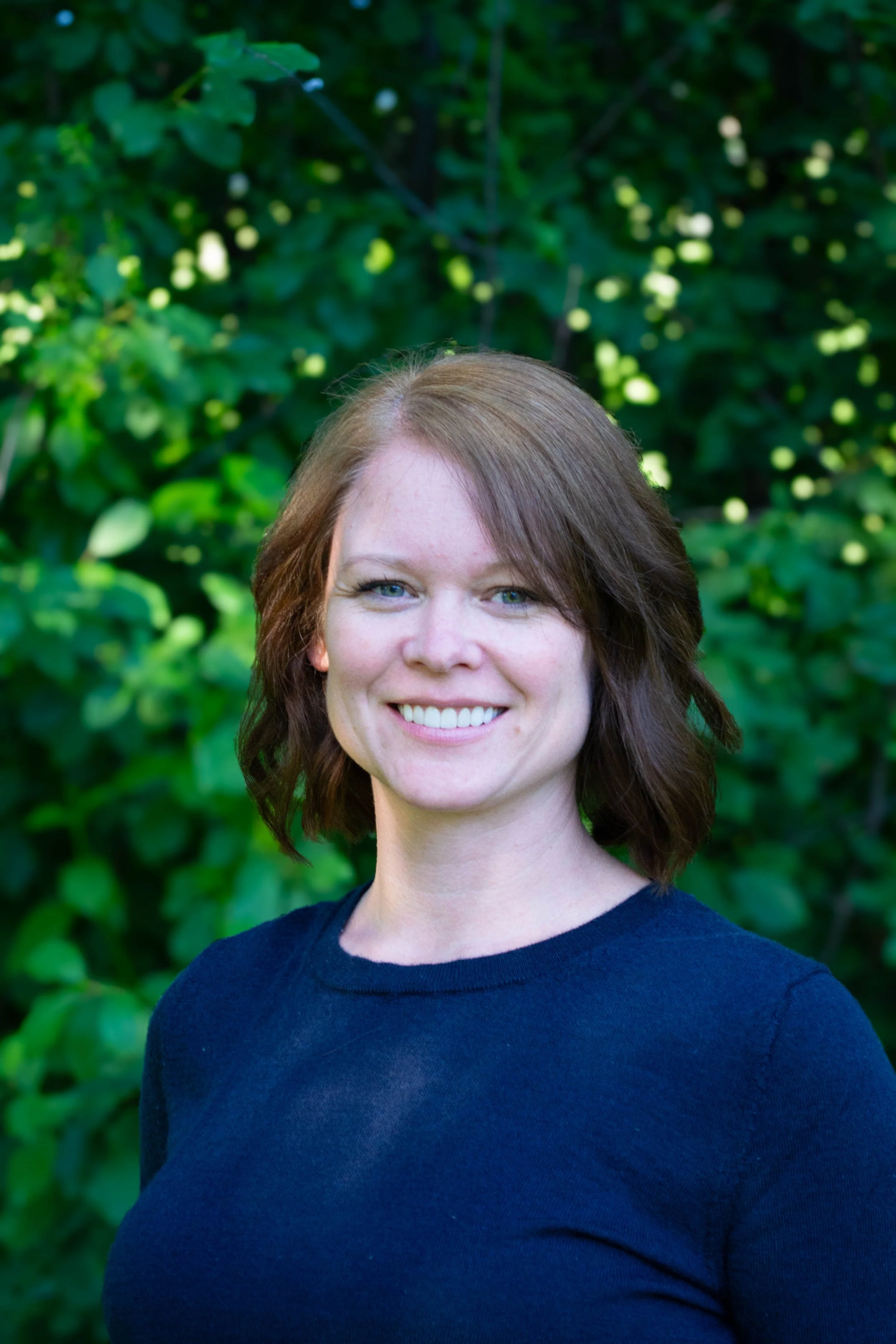 A portrait photograph of a smiling woman with light skin and reddish-brown hair, wearing a navy blue top, standing outdoors with green leafy foliage in the background.