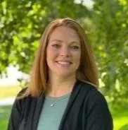A woman with shoulder-length reddish hair smiling outdoors in front of green trees.