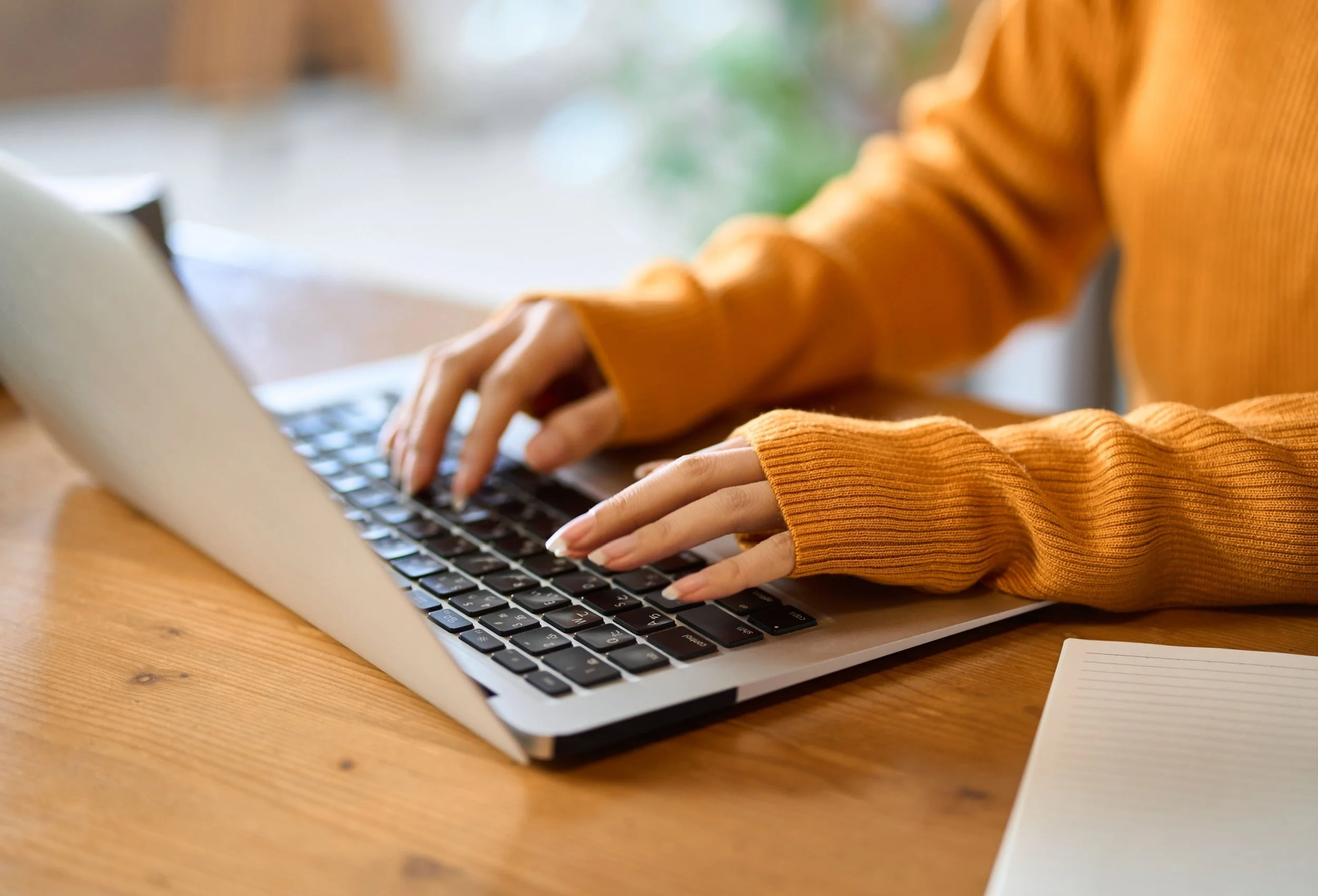Person typing on a laptop keyboard, wearing an orange sweater, on a wooden table with a notepad nearby.