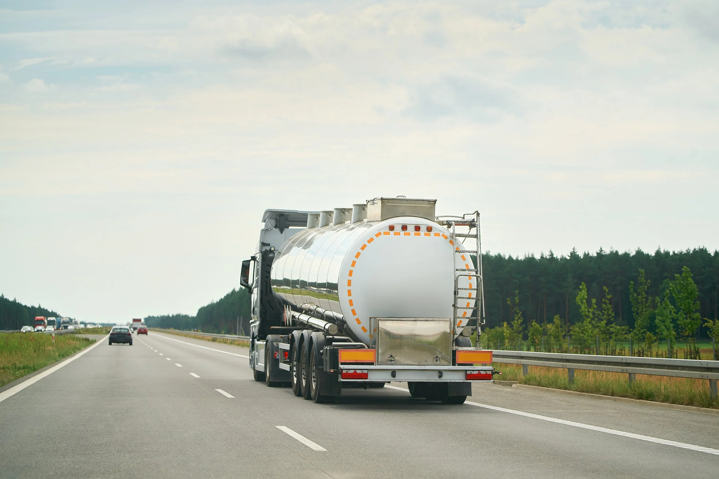 A white tanker truck driving on a highway with cars behind it and a line of trees in the background.