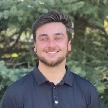 A young man with brown hair and a beard smiling outdoors, wearing a blue sweater and a collared shirt.
