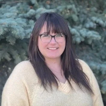 A woman with glasses and long dark hair smiling outdoors in front of evergreen trees