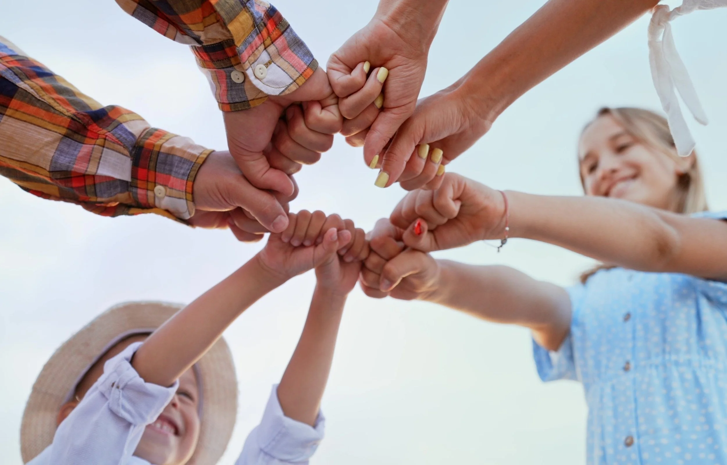 Child wearing a sunhat and white shirt smiling while and holding hands with other children in a circle, outdoors, under a cloudy sky.