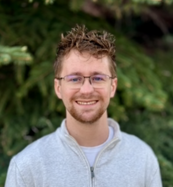 A young man with glasses, light brown hair, and a beard, smiling outdoors in front of lush green foliage.