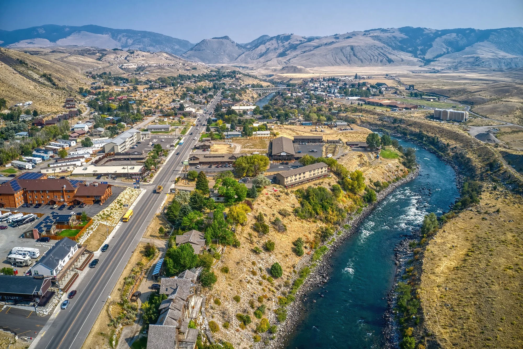 Aerial view of a small town with houses, commercial buildings, and a bridge over a river, surrounded by dry mountainous terrain with sparse vegetation.