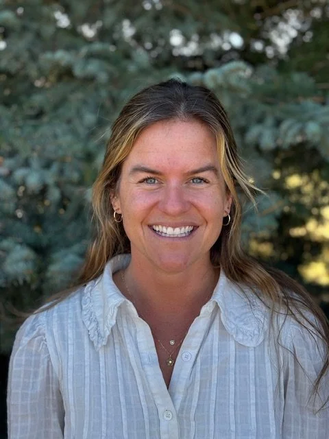 A woman with long hair smiling outdoors in front of green trees, wearing a white collared shirt.