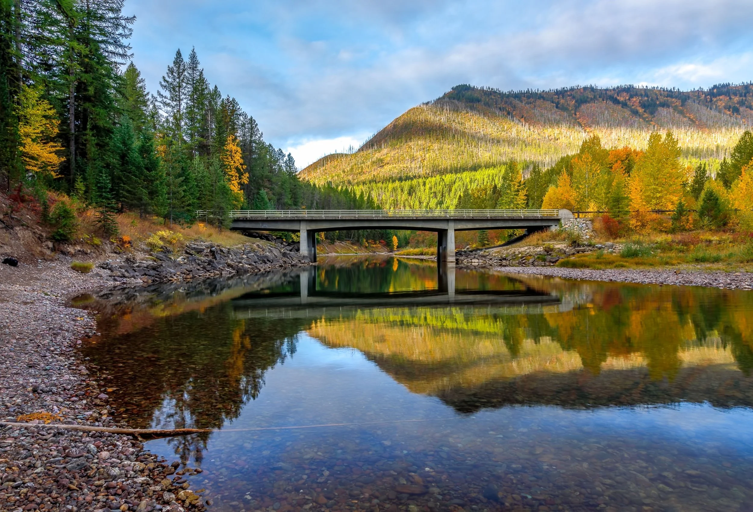 A serene landscape of a river reflecting a forested hillside during fall with colorful leaves, a bridge crossing over the river, and hills in the background under a partly cloudy sky.