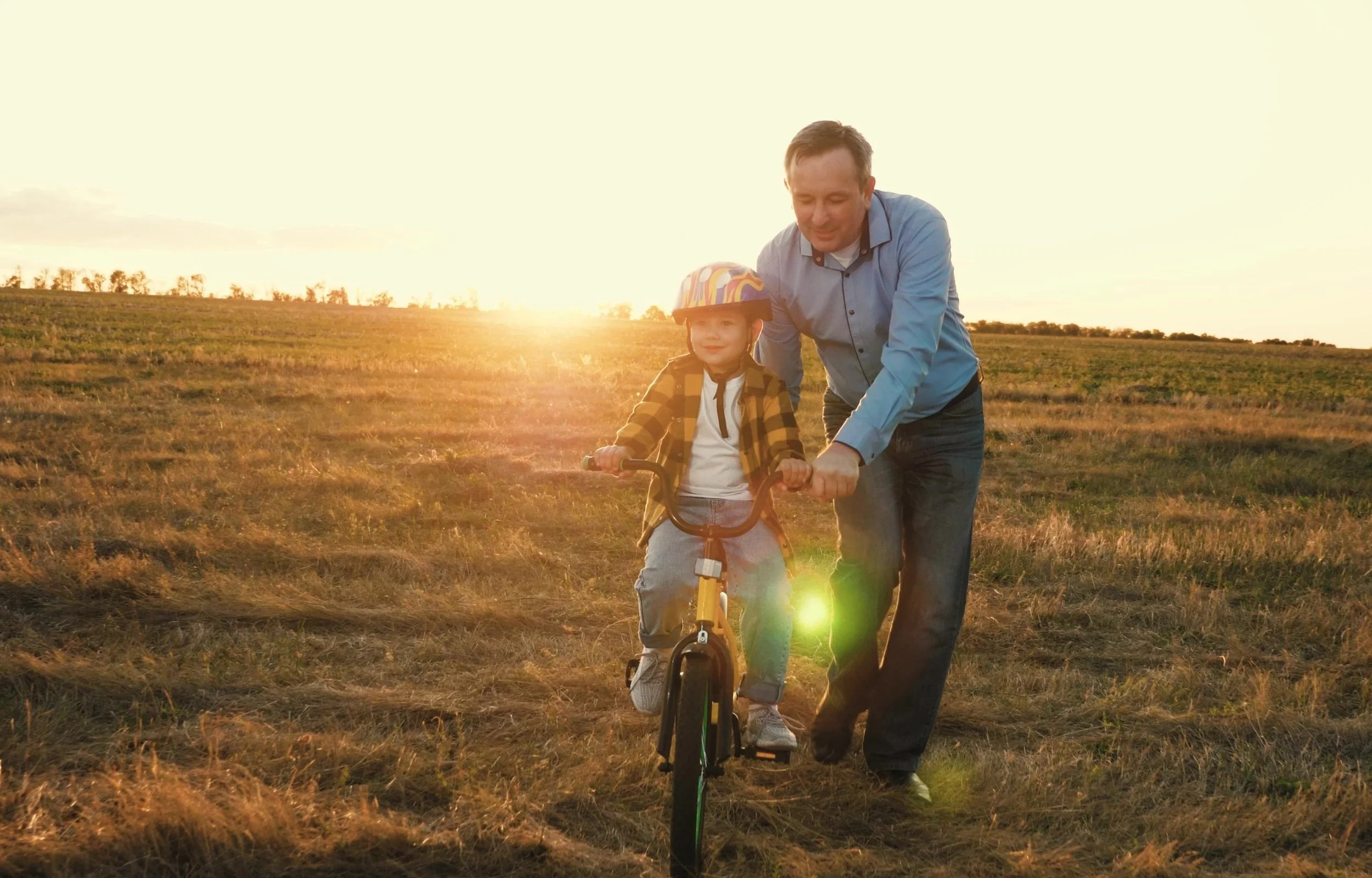 A father and son riding a bicycle together on a grassy field during sunset, with the son wearing a colorful helmet and plaid shirt.