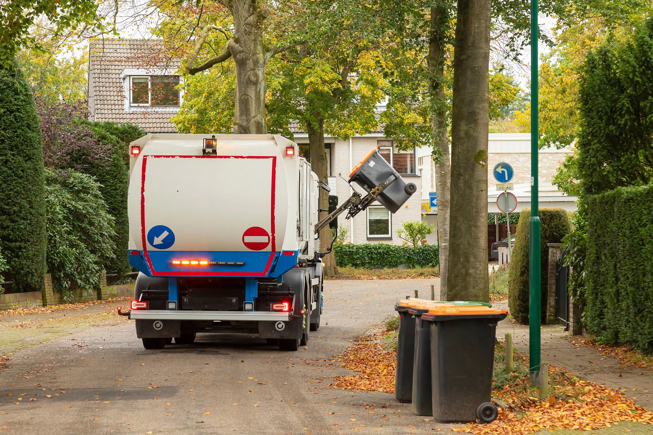 A garbage collection truck cleaning the street in a residential neighborhood with trees, houses, and trash bins along the sidewalk.