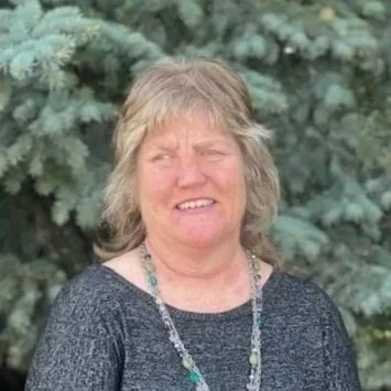 A middle-aged woman with blonde hair standing outdoors in front of pine trees, wearing a dark sweater and a beaded necklace.