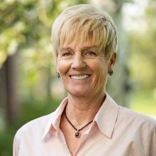 A smiling woman with short blonde hair outdoors, wearing a light-colored blouse and a necklace.