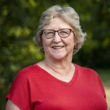 Smiling elderly woman with gray hair and glasses, wearing a red shirt, outdoors in a park.
