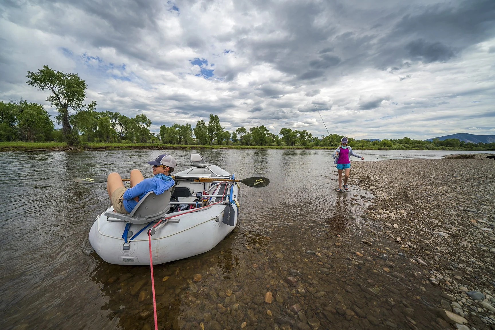 A boy sitting in an inflatable boat on a river, with a woman walking along the riverbank fishing. The sky is cloudy with patches of blue, and there are trees and mountains in the background.