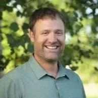 A man with brown hair and a beard smiling outdoors, wearing a green collared shirt, with trees and greenery in the background.