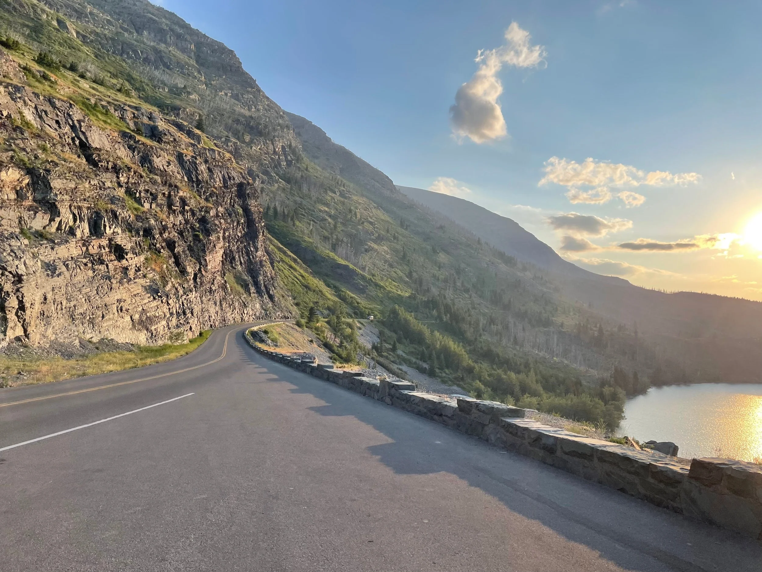 A winding mountain road runs along a lake with mountains on one side and the water on the other, under a clear sky with scattered clouds.