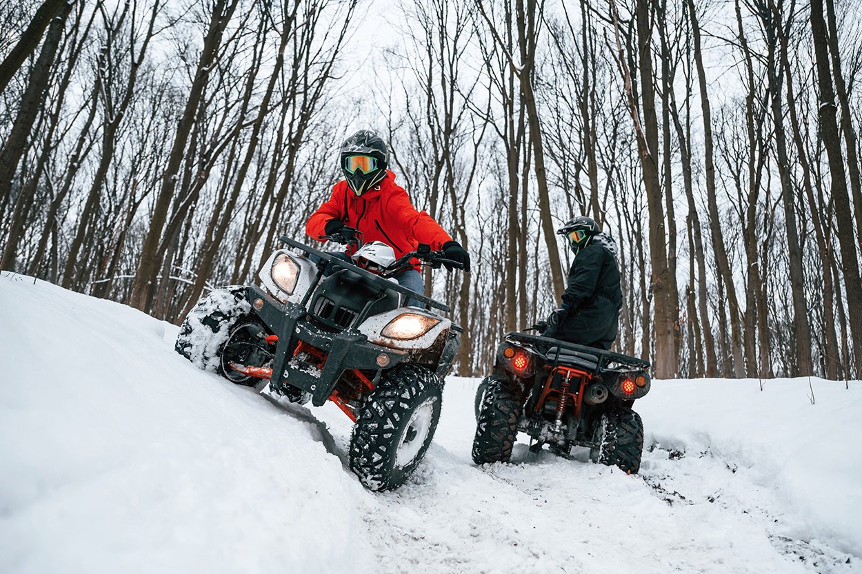 Two people riding all-terrain vehicles (ATVs) in a snowy forest, wearing winter gear and helmets. One rider is in a red jacket, and the other in a black jacket.