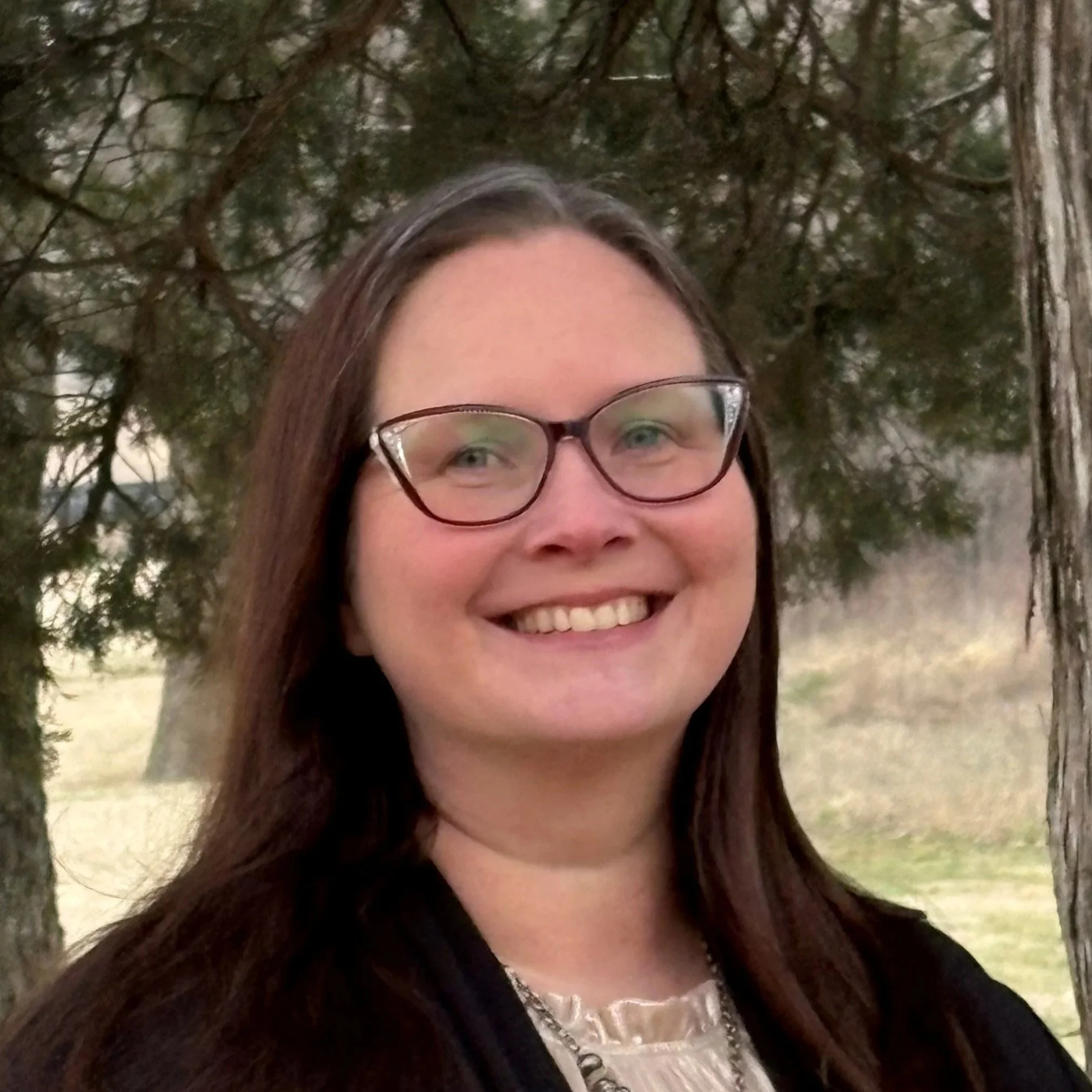 Close-up of a smiling woman with long dark hair and glasses outdoors, with trees and greenery in the background.