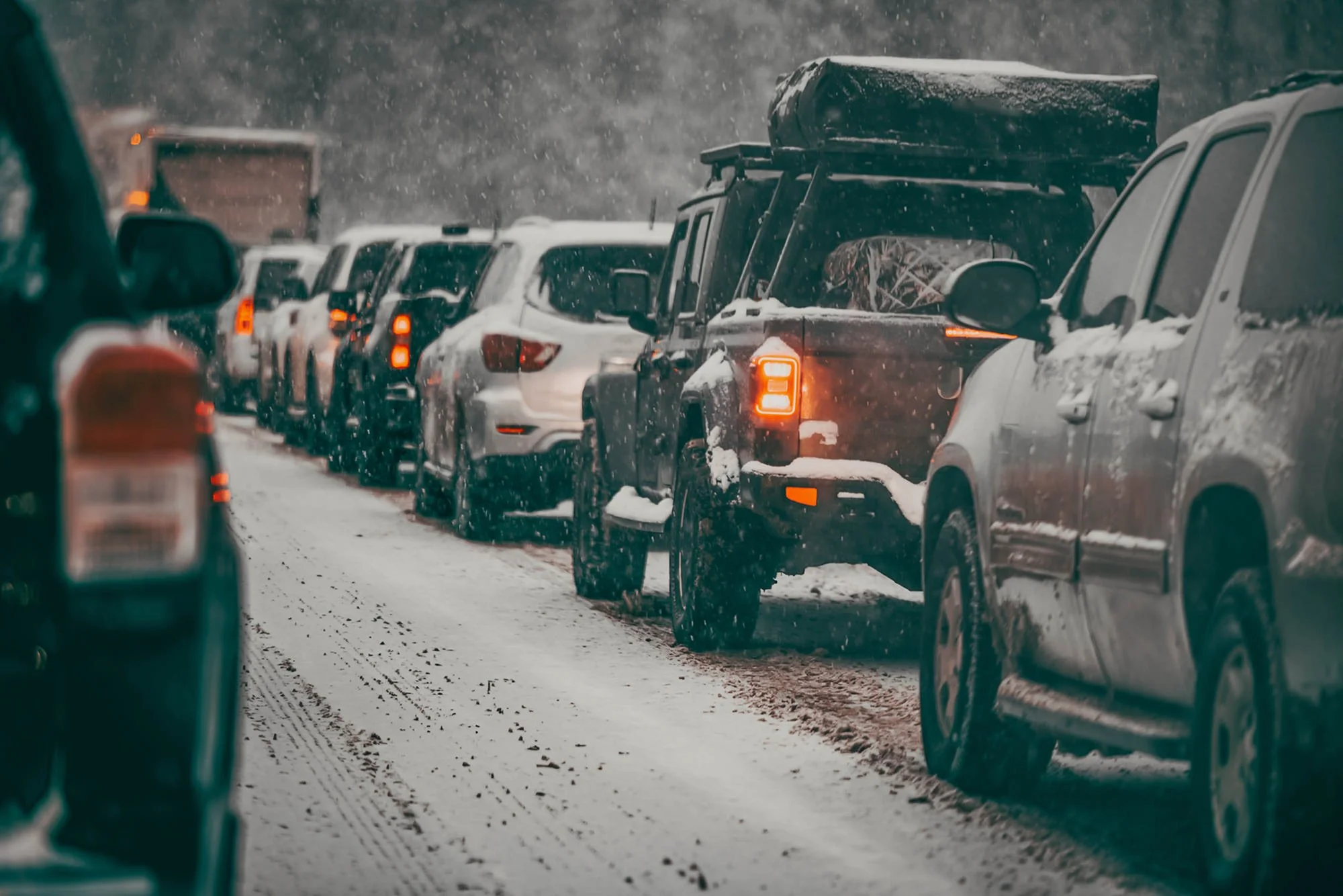 Line of vehicles on snowy road during heavy snowfall, with snow accumulating on vehicles and the ground.