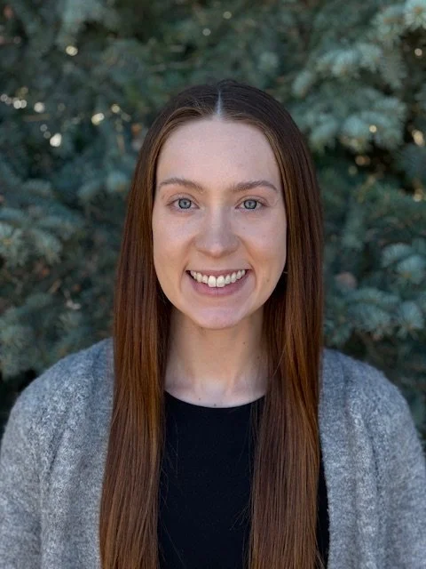 A young woman with long reddish-brown hair, smiling with white teeth, wearing a black top and gray cardigan, standing outdoors in front of green foliage.