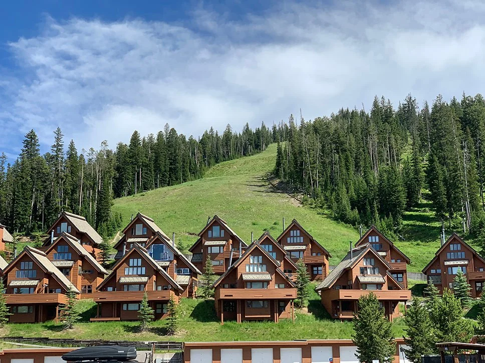 Multiple wooden A-frame houses on a hillside with green grass and pine trees, set against a background of a forested mountain and partly cloudy sky.