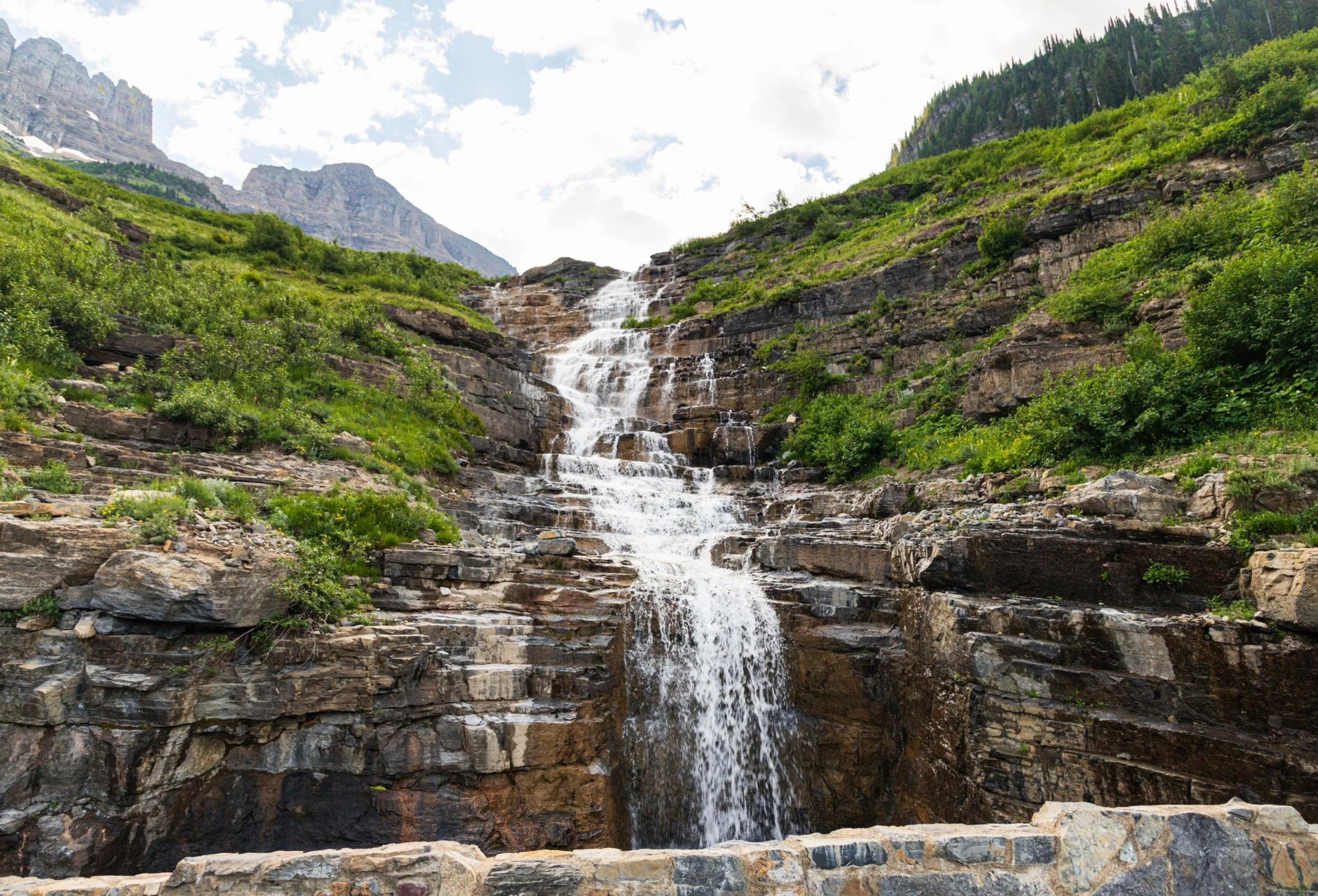 A scenic mountain landscape with a waterfall flowing over layered rocks, surrounded by green bushes and trees, with mountains and a partly cloudy sky in the background.