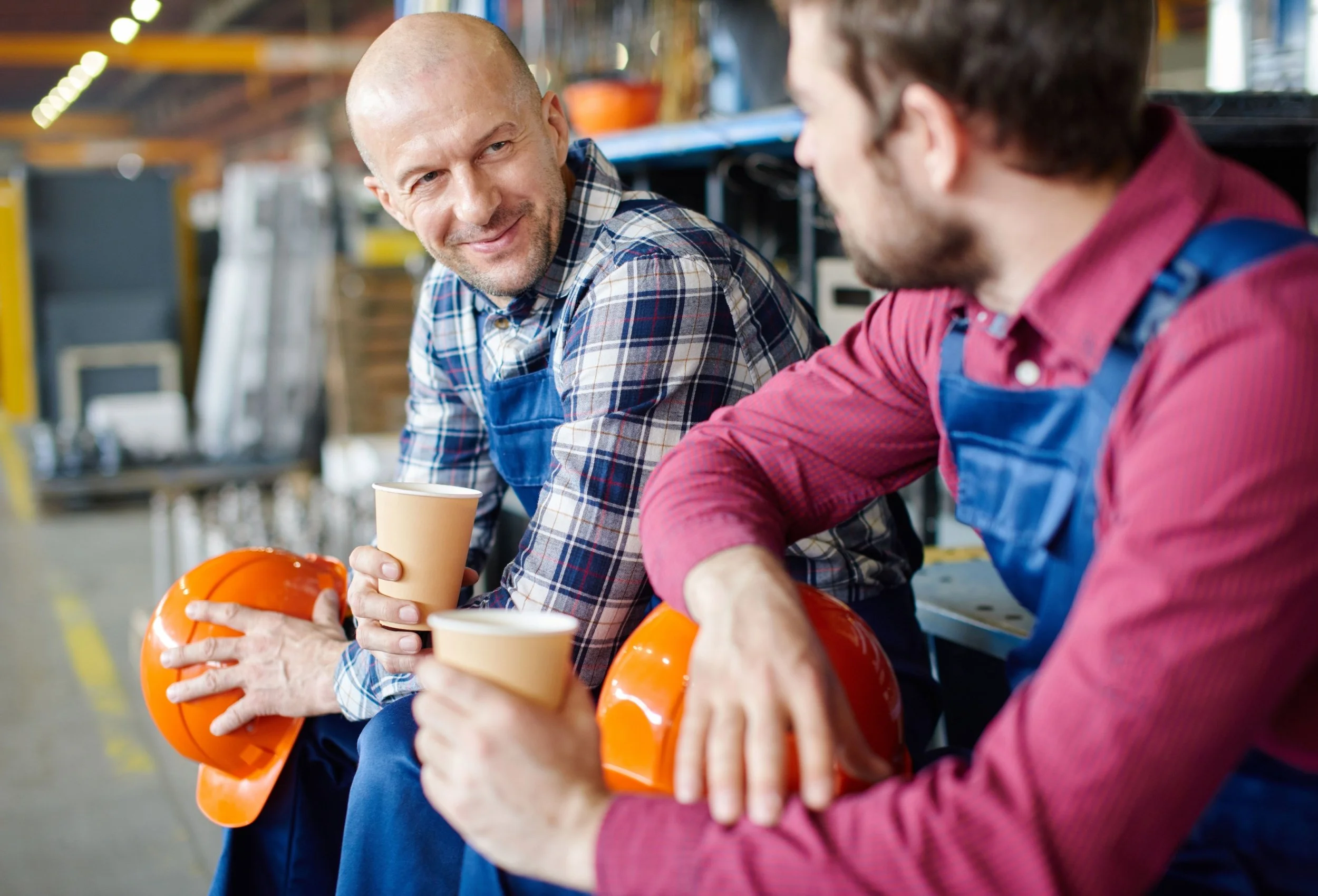 Two men with safety helmets and vests sitting on a bench in a hardware store, holding cups and talking.