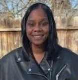 A woman with shoulder-length black hair wearing a black leather jacket, standing outdoors with a wooden fence and trees in the background.